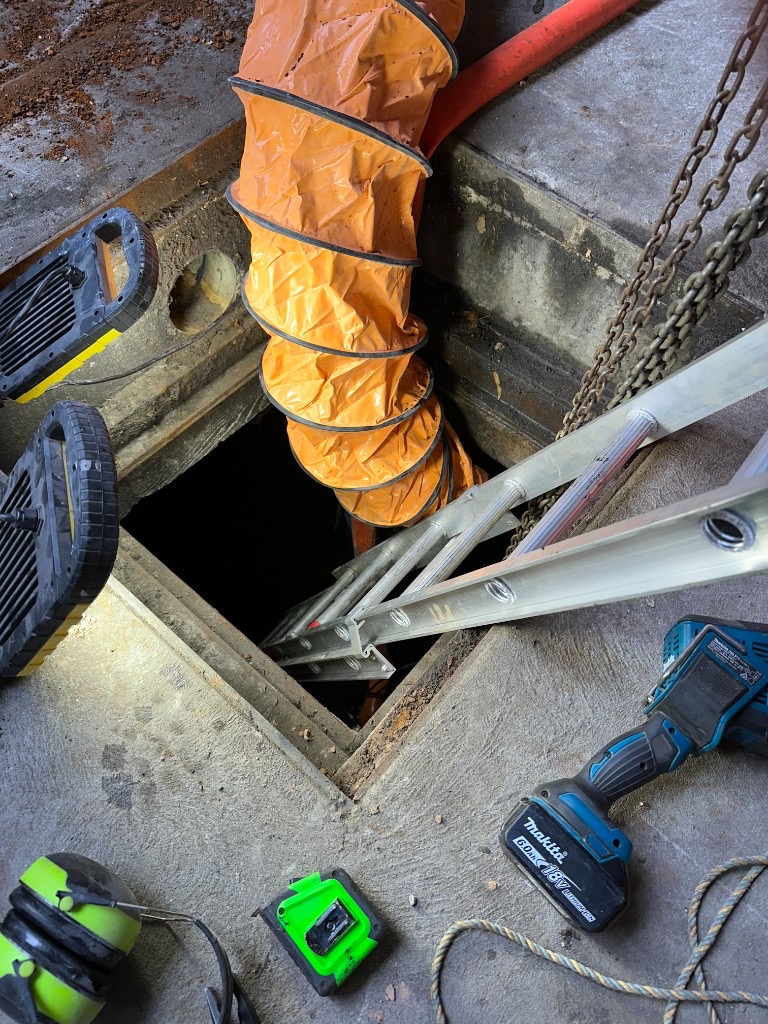 Confined space entry setup into the Queen Victoria Apartments pump station pit, with orange ventilation duct, access ladder, work lights and tools on the concrete floor